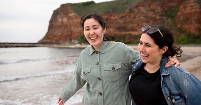 Two women laughing and enjoying a walk on the beach with cliffs in the background.