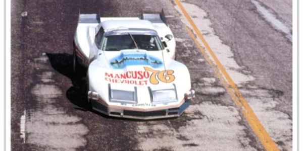 A vintage white race car with Mancuso Chevrolet branding on a weathered track.