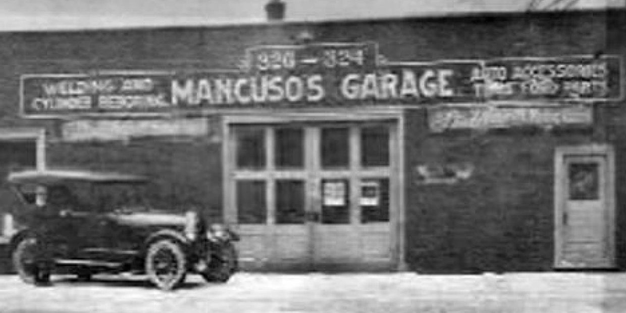 Black and white photo of Mancuso's Garage with vintage car parked outside.