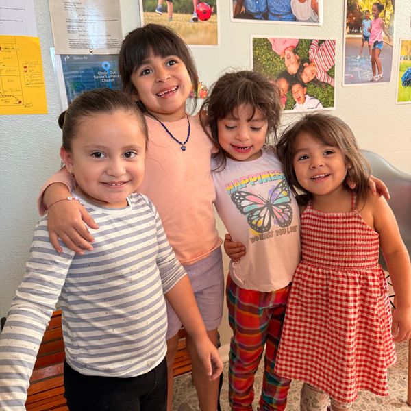 Four young girls smiling and hugging indoors with photos on the wall behind them.
