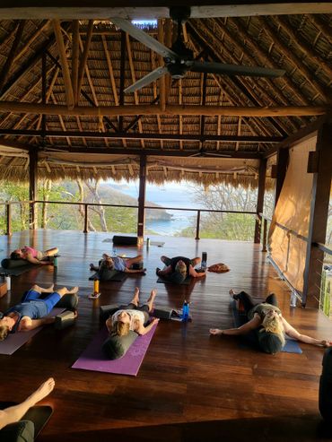 People relaxing on yoga mats in a serene open-air wooden pavilion with ocean views.