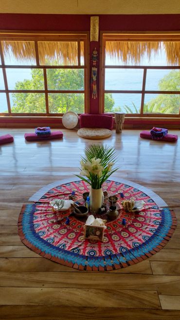Meditative space with colorful mandala cloth, flowers, and cushions by large windows overlooking trees and sea.