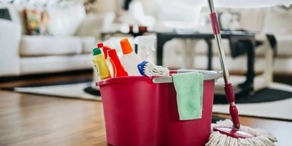 A red cleaning bucket with various cleaning supplies and a mop on a wooden floor.