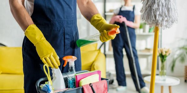Two people in blue aprons and yellow gloves preparing cleaning supplies indoors.