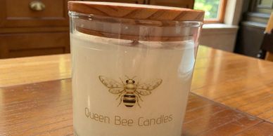 A Queen Bee Candles jar with a wooden lid on a wooden table.