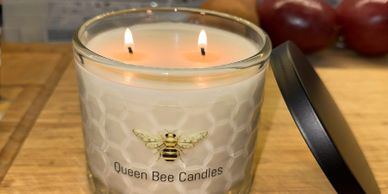 A lit Queen Bee Candles jar with two flames on a wooden surface.