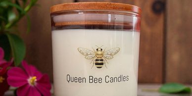 A white candle in a glass jar labeled 'Queen Bee Candles' with a bee illustration and wooden lid.