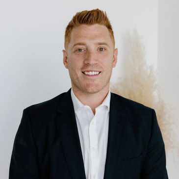 Smiling man in a black suit and white shirt against a neutral background.