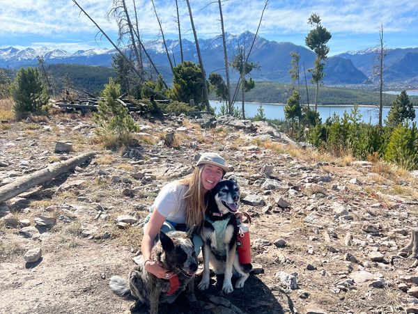 A woman posing with two dogs on a rocky trail with mountains and a lake in the background.
