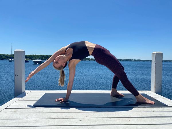Woman doing yoga backbend on a dock by the water under a clear blue sky.