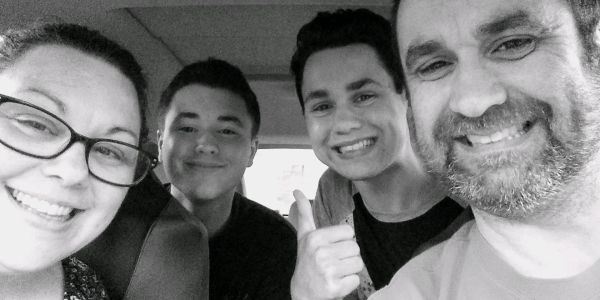 A happy family of four smiling inside a car, with one giving a thumbs-up.