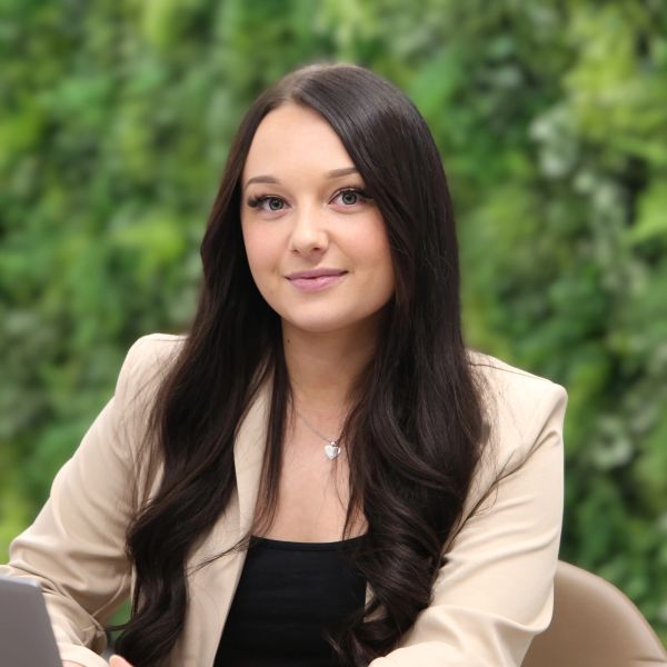 Young woman with long dark hair wearing a beige blazer, smiling gently.