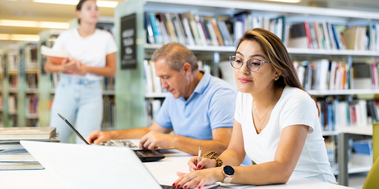 Image of 3 people in a library, with two sitting at a table on laptops and 1 person standing. 