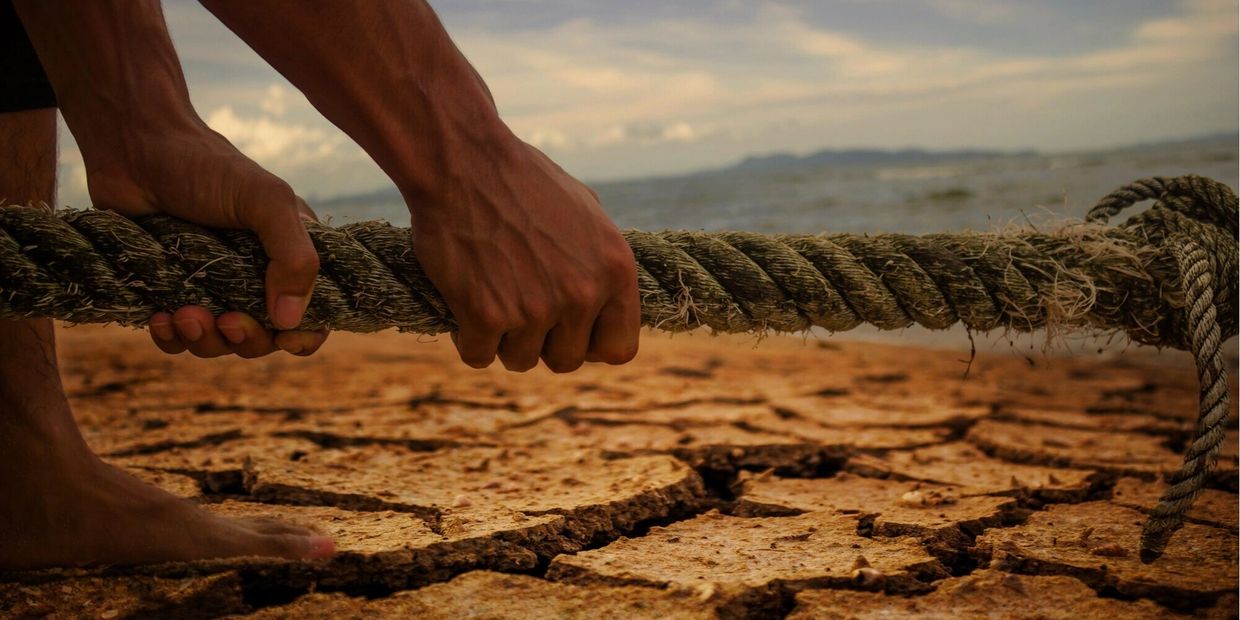 Hands gripping a thick rope on cracked dry soil near water.