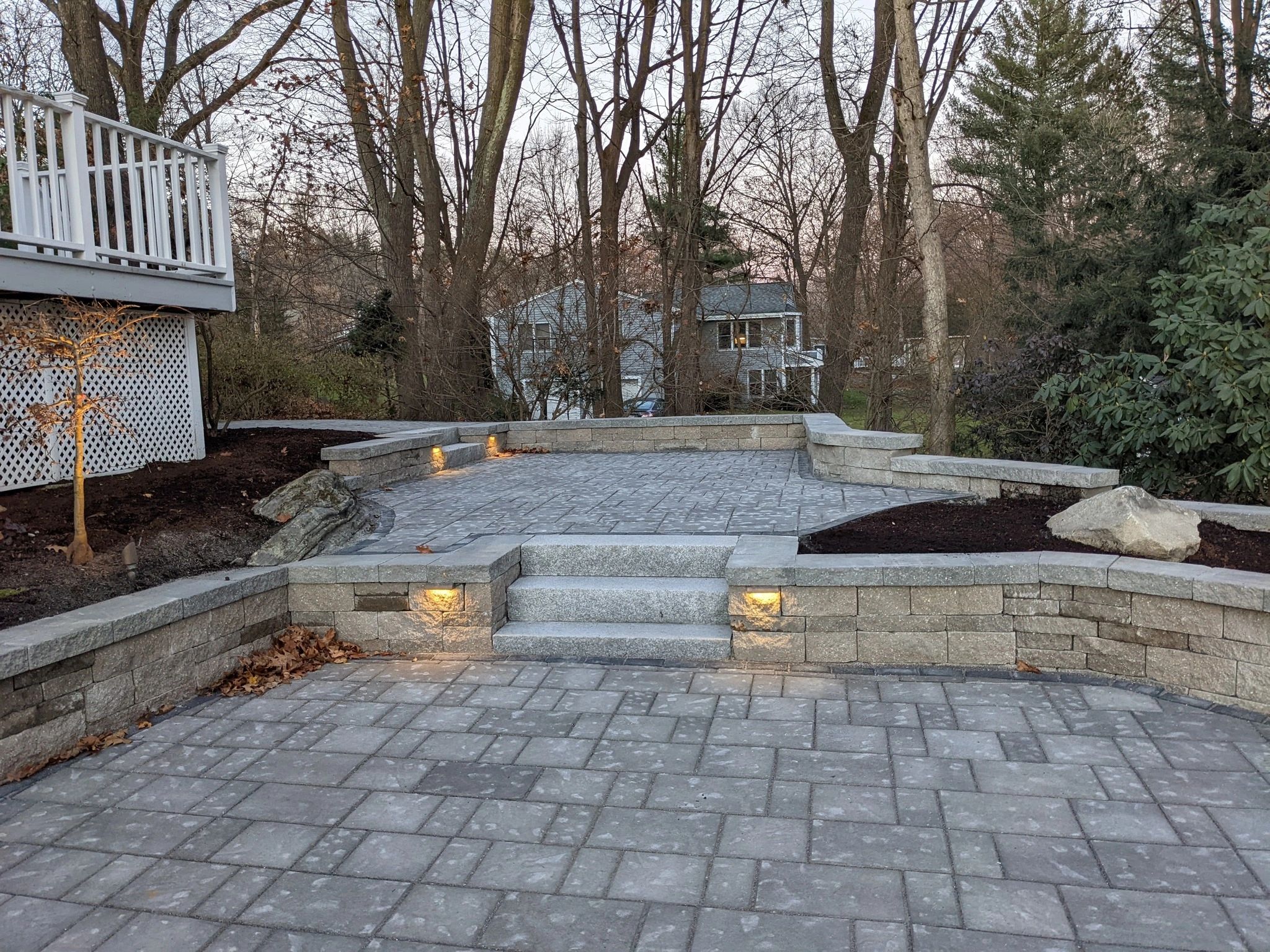 Stone patio with steps, built-in seating, and ground lights in a wooded backyard