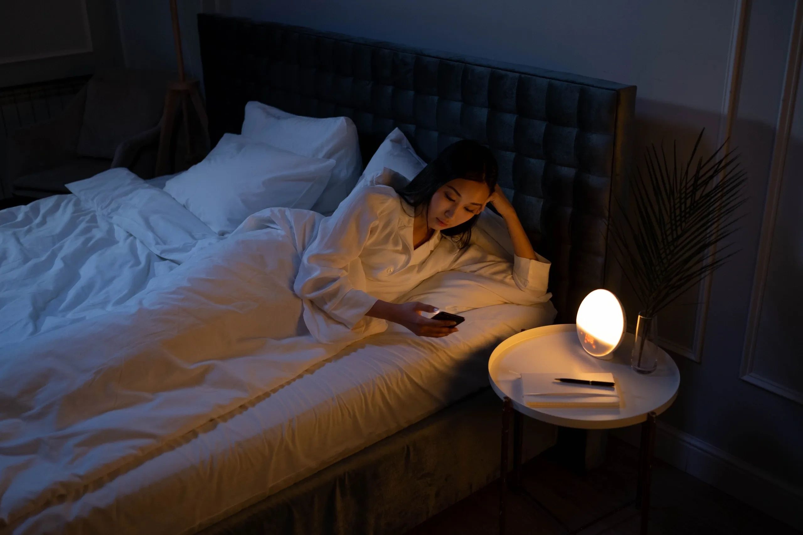 Woman in white pajamas on bed using phone in soft light.