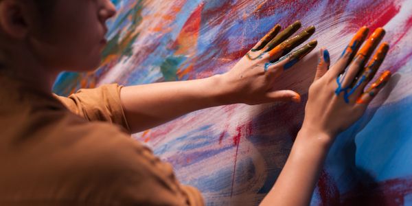 A teenaged girl in a yellow shirt, uses her hands to paint on a large canvas. 