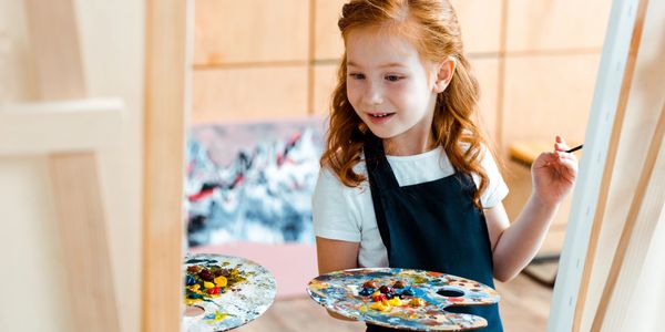 A young red haired girl smiles at her art work, holding her paint brush and pallet.