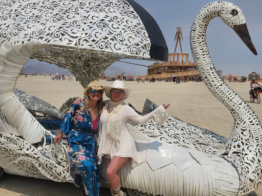 Two women pose happily beside a large ornate swan sculpture in a desert festival setting.
