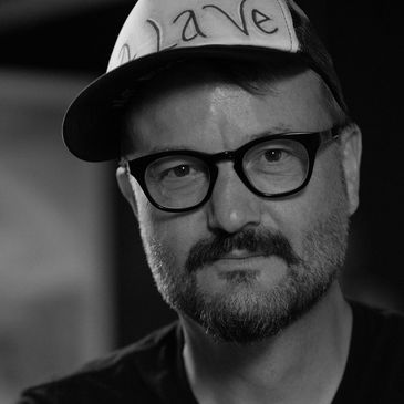 Black and white portrait of a man wearing glasses and a cap with 'Wave' written on it.