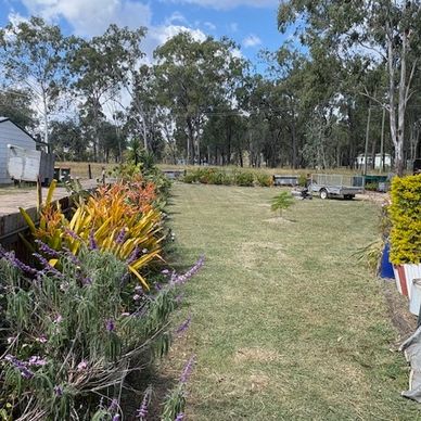 A spacious backyard with flowers, grass, and trees under a blue sky.