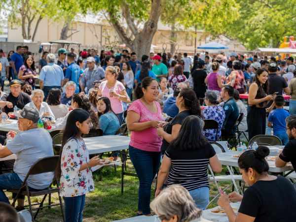 A lively outdoor community gathering with people eating and socializing under trees.