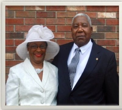 Elderly couple dressed formally, posing in front of a brick wall.