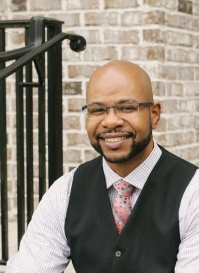 Smiling man in glasses and vest sitting outdoors.