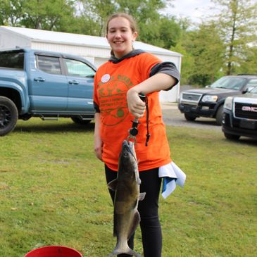 Smiling girl holding a large fish outdoors beside parked trucks.