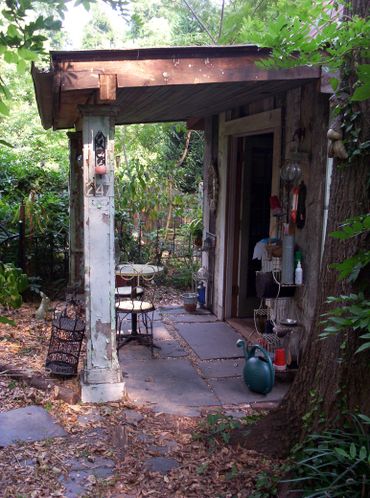 Rustic porch with vintage chairs and garden tools surrounded by lush greenery.