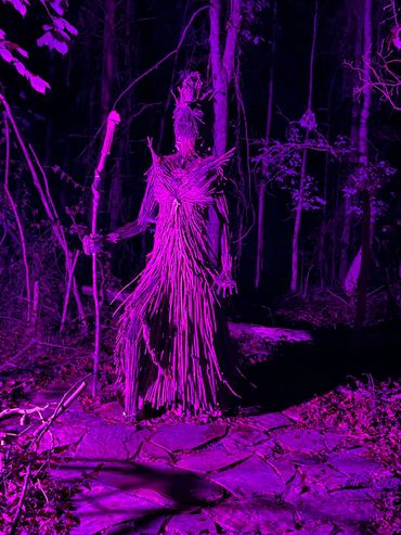 A tall figure made of sticks stands in a dark forest glowing purple.