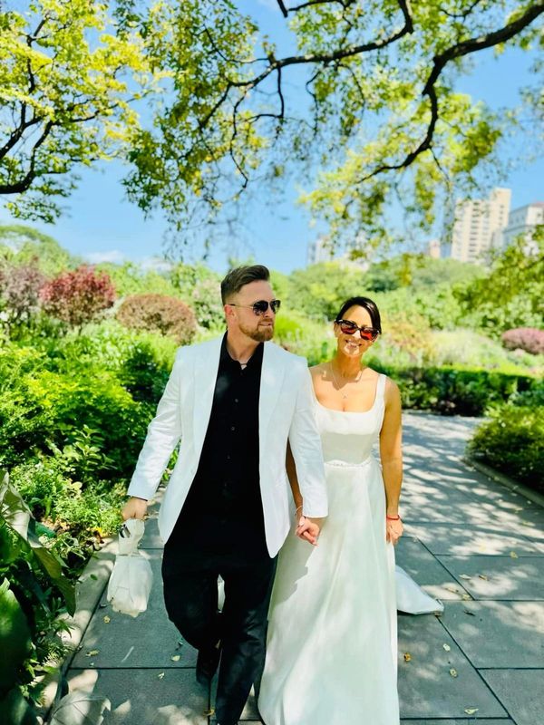 Couple dressed in white walking hand-in-hand through a sunny park.