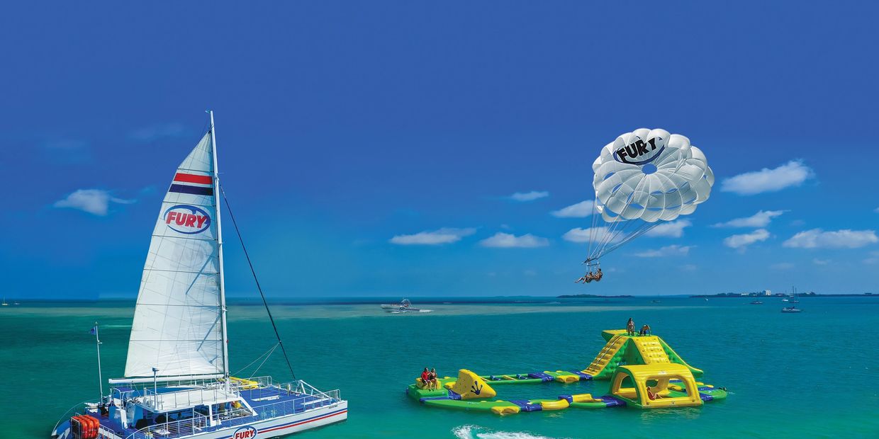 A white sailboat next to parasailers, Jetski, banana boat, and inflatable ocean water park