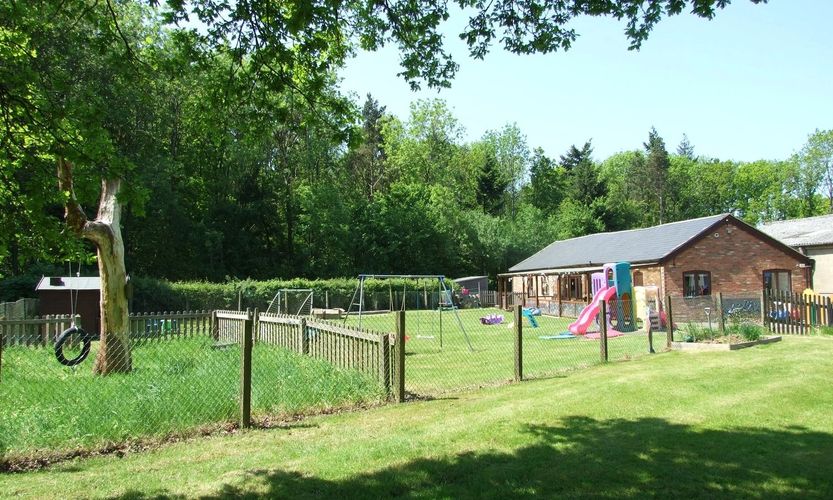 A fenced playground with swings and slides next to a brick building surrounded by trees.