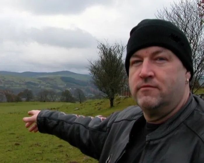 Man in black beanie and jacket pointing towards a grassy landscape with hills.