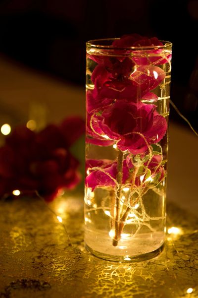 A tall glass vase with red flowers illuminated by warm fairy lights.