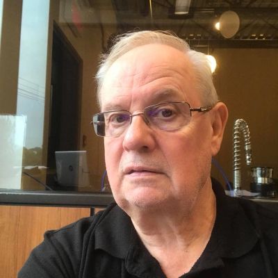 An elderly man with glasses and white hair sitting indoors.