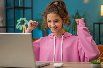 A happy girl in a pink hoodie celebrating while using a laptop.