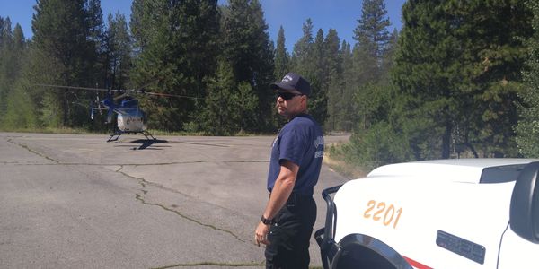 A man in uniform near a helicopter and a white vehicle in a forested area.