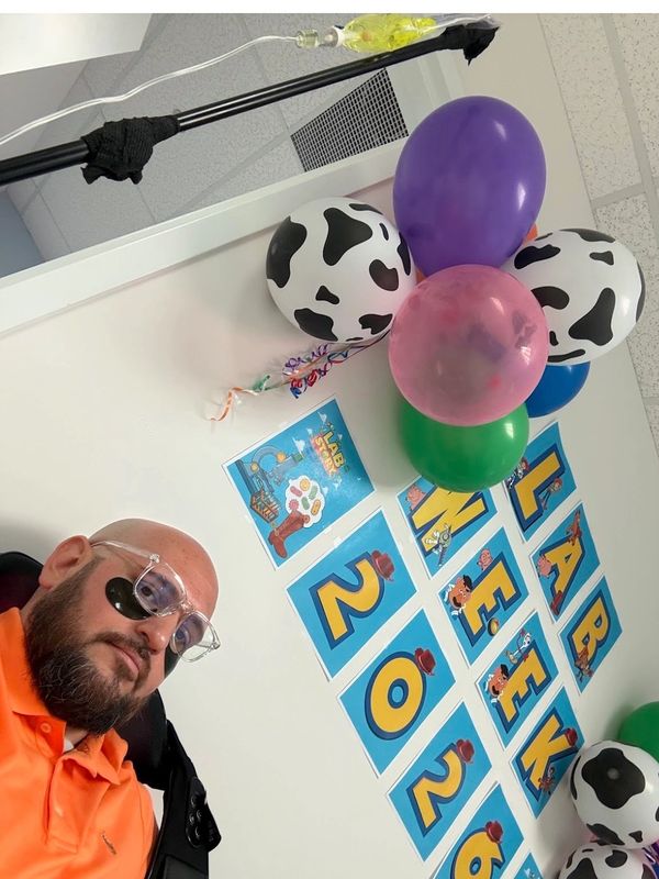 Man with eye patch in orange shirt near festive balloons and a lab week 2023 sign.