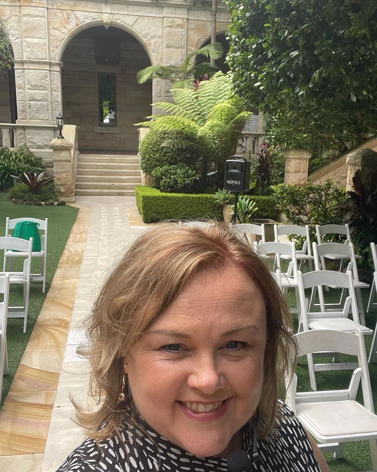 Julie Dominish smiling in an outdoor garden setting with chairs and stone architecture.