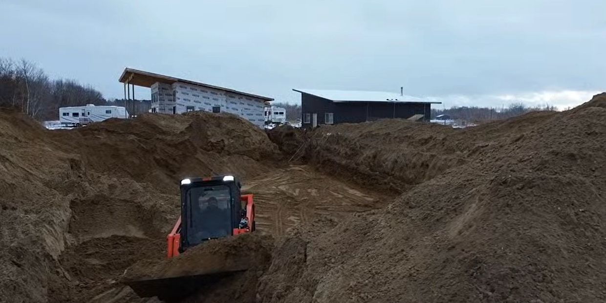 A skid steer loader moves dirt in a large excavation site near buildings.