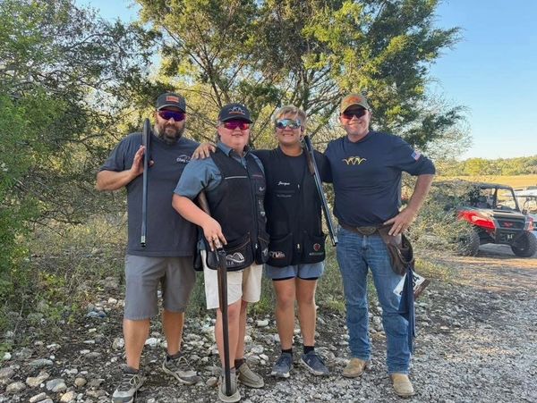 Four people posing with shotguns outdoors, dressed for shooting sports.