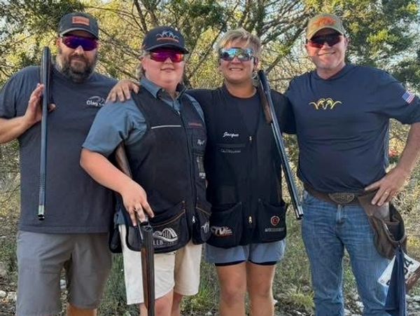 Four people posing with shotguns outdoors, dressed for shooting sports.