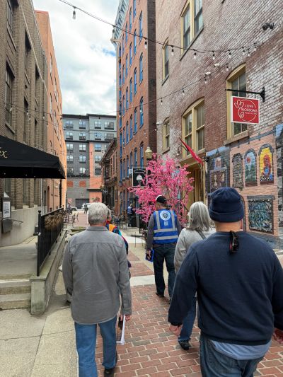 People walking down a narrow brick alley with art and a pink tree.