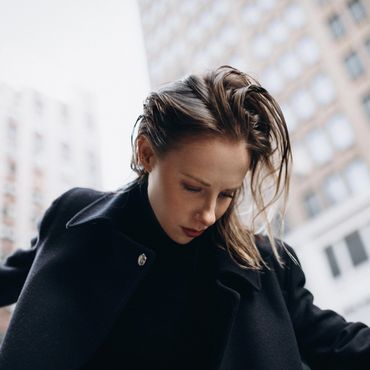 A woman in a black coat looks down against a city backdrop.