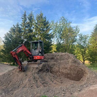 Small excavator digging a large hole in the soil outdoors.