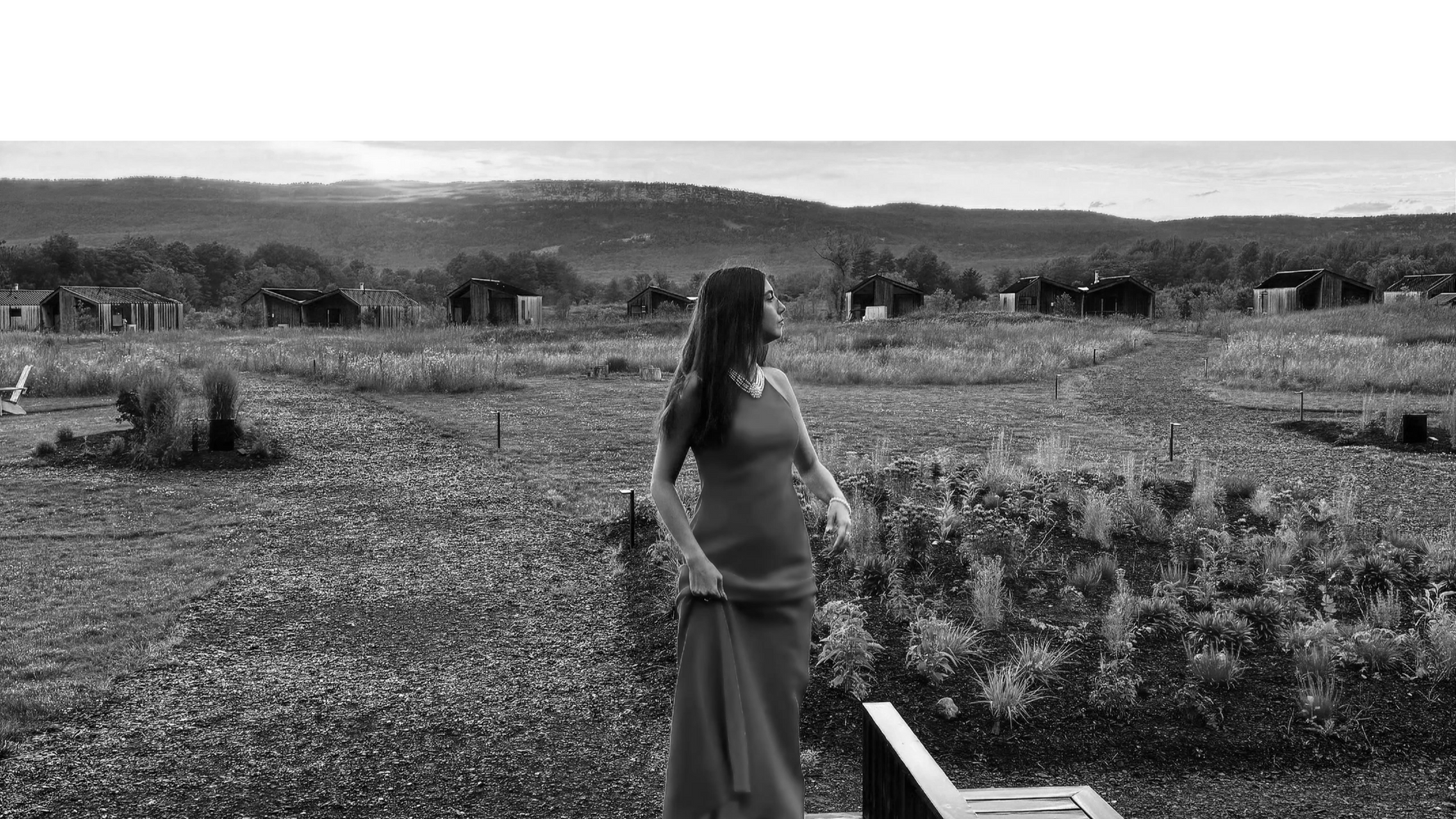 A woman in a red dress stands on a pathway in a scenic countryside with wooden cabins.