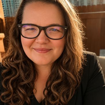 Smiling woman with glasses and curly hair in warm sunlight.