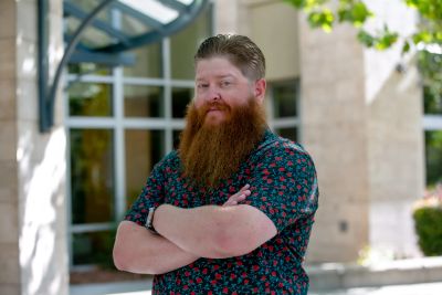 Man with a red beard and floral shirt standing with arms crossed.